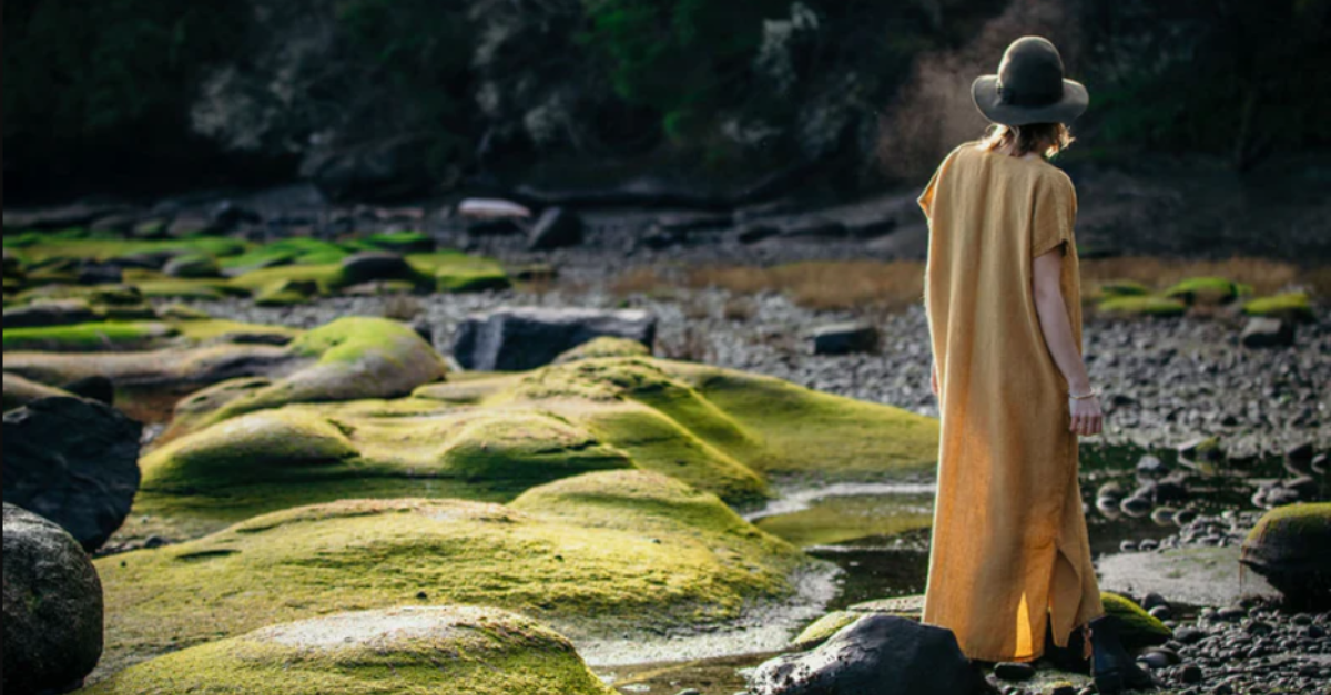 Model wearing a long yellow dress with a brown hat on standing on the rocky beach and moss covered rocks | Elizabeth Homestead