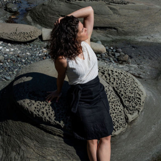 Aprons | Elizabeth Homestead (Individual sitting on a rock on the beach holding their hair back feeling the sun on their face modeling a black apron from Elizabeth Homestead)